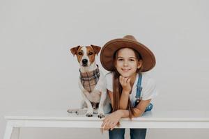 tevreden mooi meisje draagt hoed, leunt naar witte tafel, poseert in de buurt van rashond, geniet van vrije tijd samen, gaat wandelen, rust thuis. kinderen, geluk, dieren, lifestyle concept foto