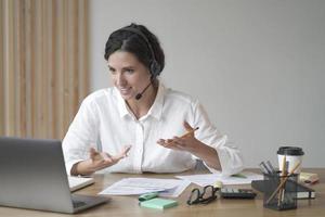 lachende vrouw online tutor zittend aan bureau in headset en praten via video-oproep op laptopcomputer foto