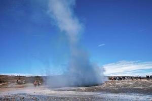 ontdekkingsreizigers kijken naar de uitbarsting van de Strokkur-geiser in de vallei tegen de blauwe lucht foto