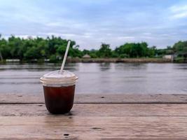 iced americano koffie in afhaalmaaltijden transparante plastic beker op houten tafel met vers kalm uitzicht op de rivier foto