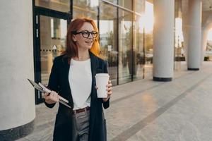 vrouwelijk model houdt touchpad-notebook vast en kopje afhaalkoffie poseert buiten tijdens zonnige dag foto