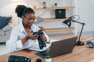 bij de tafel zitten. jonge Afro-Amerikaanse vrouw in wit overhemd is thuis foto