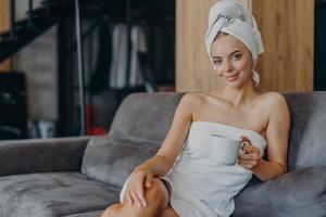 ontspannen mooie jonge vrouw heeft een gezonde huid draagt een zachte handdoek op het hoofd en rond het naakte lichaam houdt een mok koffie poseert op de bank met aromatische drank geniet van het weekend heeft een goed verzorgde teint foto