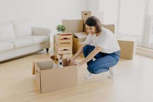 nieuw huis, verhuisdag en verhuisconcept. Positieve brunette vrouw speelt met rashond in kartonnen container, dozen met spullen uitpakken, poseren in ruime woonkamer met comfortabele bank foto