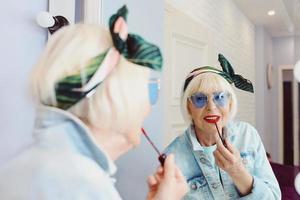 lachende oudere senior stijlvolle vrouw in blauwe zonnebril en spijkerjasje met rode lippenstift bij de spiegel in stijlvol loft interieur. stijl, mode, make-up, anti-age concept foto