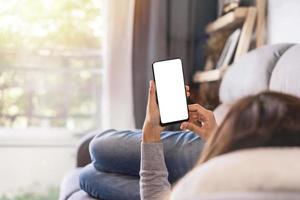 jonge vrouw die op de bank in de woonkamer ligt en thuis mobiele telefoon gebruikt foto