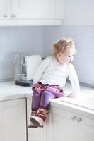 schattig peuter meisje zit in witte keuken op het aanrecht foto