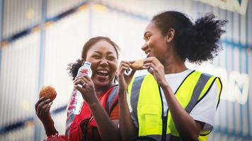 glimlach twee Afro-Amerikaanse vrouw voorman werknemer of vrouw onderhoudsingenieur in reflecterend vest veiligheidsvest gaat zitten op oude vrachtwagen ontspannen, eet brood, snacks en water tijdens de brunchpauze