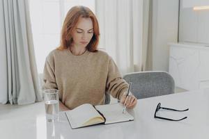 serieuze roodharige jonge vrouw maakt aantekeningen in notitieblok zit aan tafel foto