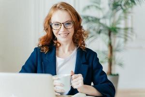 Vrolijke roodharige vrouw bereidt sollicitatiebrief voor, zit voor geopende laptop, surft op internet en zoekt naar vacatures, houdt mok met warme drank vast, draagt bril en formele outfit foto