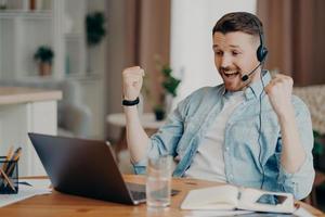 dolblij triomfantelijk volwassen man balt vuisten viert succes kijkt aandachtig naar laptop scherm draagt headset heeft zakelijke bijeenkomst online poseert op desktop tegen wazige huisachtergrond foto