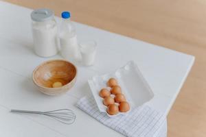 ingrediënten voor het bakken op witte tafel. eieren, melk en bloem in containers. klopper voor het kloppen en maken van deeg. proces van bakken. tijd om te koken foto