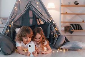 smartphone gebruiken. twee kleine meisjes zitten samen in de tent in de huiskamer foto