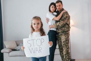 meisje met banner met stop oorlogstekst erop. soldaat in uniform is thuis met zijn vrouw en dochter foto