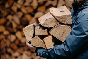 onherkenbare man draagt een hoop hout voor het maken van vuur gekleed in een jas. anonieme man draagt brandhout in huis foto