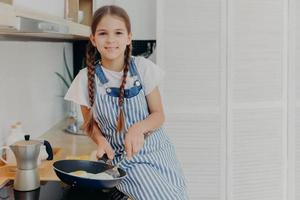 binnenopname van een prettig uitziend zevenjarig meisje dat bezig is met het bakken van eieren, houdt de pan vast en zit bij het fornuis, gekleed in een schort, kijkt positief naar de camera, schept op dat ze kan koken, poseert in de moderne keuken foto