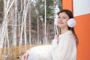 tiener meisje zittend op een staicase van een bos modern huis en camera kijken. meisje met witte pluizige oorbeschermers en casual pullover ontspannen buiten in de buurt van haar huis. mooi meisje portret foto