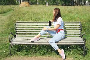 jonge mooie vrouw zittend op een bankje en genieten van aangenaam weer. zijaanzicht. vrouw die opzij kijkt en een smartphone vasthoudt. alleen reizen en lokaal reisconcept foto