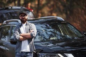 met gekruiste armen. bebaarde man in de buurt van zijn gloednieuwe zwarte auto in het bos. vakanties concept foto