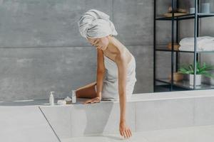 spa-, wellness- en huidverzorgingsconcept. serieuze vrouw gewikkeld in badhanddoek, gebruikt cosmetische producten om voor het lichaam te zorgen, in bad te gaan, poseert op badkuip. ontspannen vrouw thuis. spa meisje foto