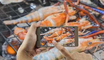 man gebruikt mobiele telefoon en maakt foto van gegrilde garnalen boven hete houtskool