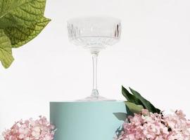 kristal cocktailglas op blauw podium onder roze bloemen en groene bladeren. vakantieselectie, bardrankjes, feestelijke decoratie. bruiloft glaswerk foto