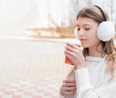 tiener meisje met gesloten ogen koffie drinken om te gaan. kopieer ruimte voor tekst. witte pullover en stijlvolle oorkappen. schoolmeisje ontspannen en wandelen in een park foto