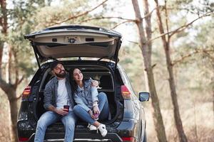 vooraanzicht. zittend op het achterste deel van de auto. genieten van de natuur. stel is aangekomen in het bos met hun gloednieuwe zwarte auto foto