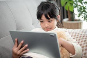 klein meisje met tablet-spel op internet, kind zittend op de bank kijkend of pratend met een vriend online, kind ontspannen in de woonkamer in de ochtend, kinderen met nieuw technologisch concept foto