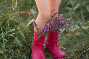 rode rubberen laarzen aan de voeten van een meisje met een boeket wilde bloemen. bloemen in een laars, zomertijd. zomer, vrijheid, natuur, platteland, groen gras in het veld foto