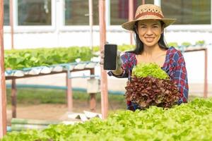 aziatische vrouw boeren werken met behulp van mobiel in groenten hydrocultuur boerderij met geluk. portret van vrouwelijke boer die de kwaliteit van groene salade groente controleert met een glimlach in de groene huisboerderij. foto