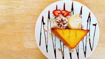 bovenaanzicht van zoete honing toast met vanille-ijs, slagroom, chocolade siroop en stuk aardbei op witte plaat en houten achtergrond foto