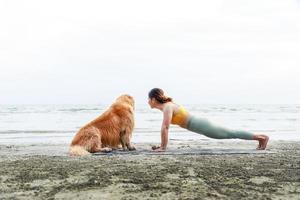 vrouw yoga-oefeningen met haar hond op het strand en kijkend naar de zee. gezond actief levensstijlconcept. ontspanning met een huisdier. foto