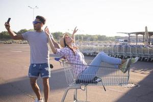 gelukkig jong stel rijden op trolley op lege parkeerplaats in winkelcentrum, hipster vriend heeft veel plezier tijdens het winkelen, verliefd paar rijden op winkelwagentje foto