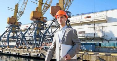 jonge mannelijke arbeider van zeehaven in helm, vrachtmanager in pak en halmet werkt buiten, kranen en zee-achtergrond foto