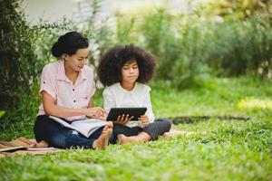 vrolijke jonge mooie moeder zit met zoon met krullend haar in het park en helpt hem tijdens het gebruik van digitale tablet tijdens het studeren foto