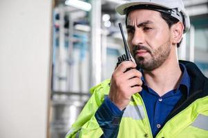 jonge mannelijke machine-inspecteur met vest en veiligheidshelm met koptelefoon die machine en sterilisatoren in waterplant controleert terwijl hij aantekeningen maakt in digitale tablet foto