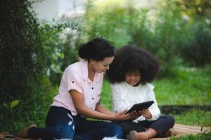 vrolijke jonge mooie moeder zit met zoon met krullend haar in het park en helpt hem tijdens het gebruik van digitale tablet tijdens het studeren foto