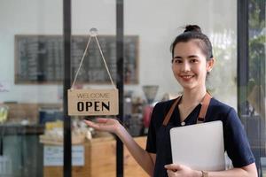 mooie aziatische jonge barista-vrouw in schort die tablet vasthoudt en voor de deur van café staat met open bord. ondernemer opstarten MKB ondernemer concept. foto