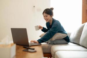 kalme jonge aziatische vrouw die werkt op een laptop die op een gezellige bank zit, een gelukkig meisje dat op internet surft of online winkelt tijdens een zonnig weekend thuis, ontspannen op de bank met de computer foto