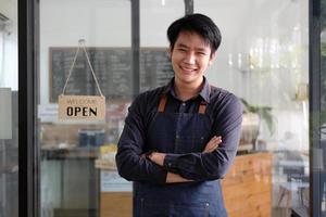 gelukkige aziatische jonge barista-man in schort die tablet vasthoudt en voor de deur van café staat met open bord. ondernemer opstarten mkb ondernemer concept foto