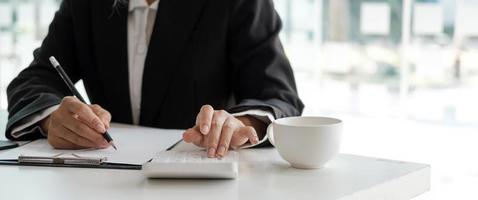 crop shot van accountant vrouw hand druk op rekenmachine en schrijven van een notitie voor het maken van financieel verslag of bedrijfswinst jaarlijks aan bureau, boekhouder baan foto