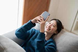 afbeelding van een glimlachende aantrekkelijke aziatische vrouw die een mobiele telefoon gebruikt terwijl ze thuis op de bank ligt foto
