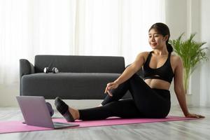 fit sportieve jonge aziatische vrouw online training oefening thuis. actieve gezonde meid geniet van sport pilates yoga fitness training op laptop computer die zich uitstrekt op yoga mat kijken naar video klasse foto