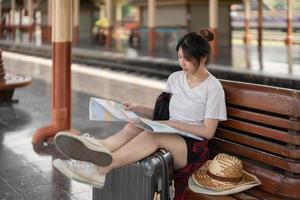 gelukkige jonge Aziatische vrouw reiziger of backpacker met behulp van kaart kiezen waar te reizen met bagage op treinstation, zomervakantie reisconcept foto