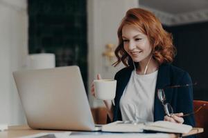 jonge vrouwelijke lerares geeft online les, praat met leerlingen via laptop, organiseert videoconferentie, geeft les aan studenten, drinkt aromatische koffie, houdt een bril in de hand, is formeel gekleed foto