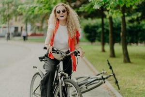 aangenaam uitziende jonge vrouw met zonnebril, rijdt op sportfiets, stopt voor pauze, geniet van schoonheid van de natuur, rustige sfeer, lacht positief, houdt beide handen aan het stuur, ontdekt nieuwe plek foto