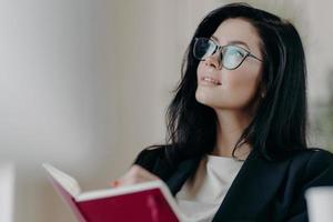 indoor shot van doordachte dromerige brunette vrouwelijke journalist werkt aan boekbespreking, houdt geopend notitieblok vast, schrijft enkele ideeën of memonotities op, maakt nieuw artikel, werkt aan project, draagt een bril. foto