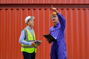 twee professionele aziatische mannelijke arbeiders in veiligheidsuniformen en veiligheidshelmen op een stalen containerachtergrond, werken op de logistieke terminal, laden controleverzendingsgoederen voor de vrachtvervoersindustrie. foto