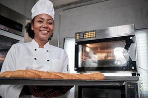 portret van een Afro-Amerikaanse vrouwelijke chef-kok in een wit kookuniform die naar de camera kijkt met een vrolijke glimlach en trots met een dienblad met stokbrood in de keuken, professionele banketbakkers, verse bakkerijbezetting. foto